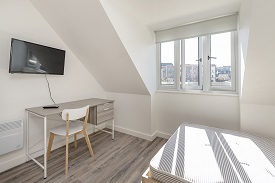 A bedroom in Old Vicarage. To the left of the frame is a desk and chair facing the wall. A television is mounted on the same wall. The room has slanted walls either side of the window. A bed is to the right of the window.